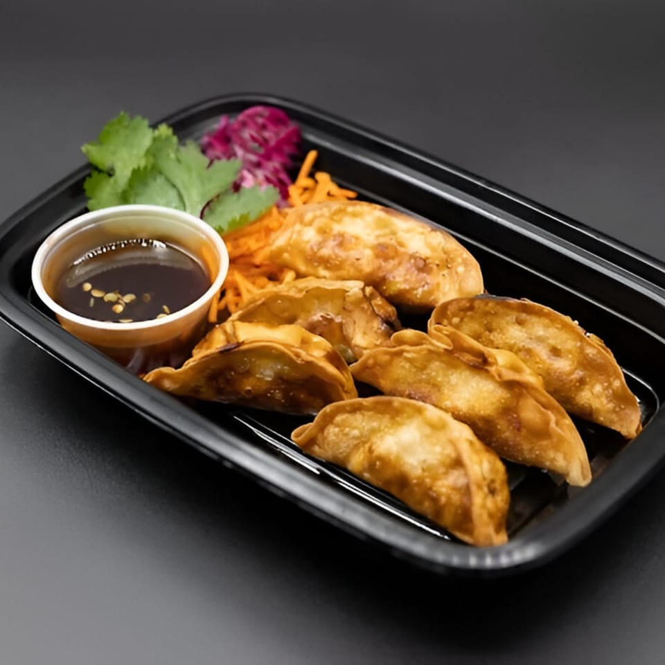 Golden-brown fried dumplings, with a side of dipping sauce, shredded carrots, and herbs in a black container.